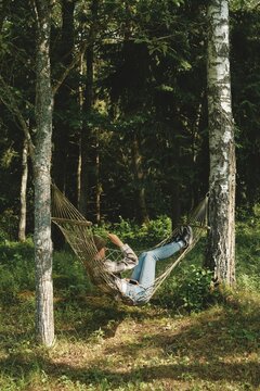 Young woman rests in hammock tied between two trees in shade in green forest during sunny day