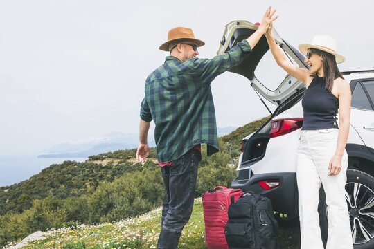 Happy Travelers tourist couple hi5 stand put luggage to white rented SUV vehicle trunk on scenic Mediterranean viewpoint. Weekend get away road trip arrival, travel preparation, planning holiday\