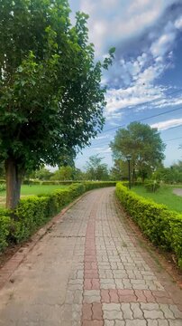 Walking Along a Curved Park Pathway Surrounded by Lush Green Trees and Beautiful Landscape &ndash; Scenic Nature Walk on Brick Track with Fresh Greenery, Blue Sky and Peaceful Outdoor Environment