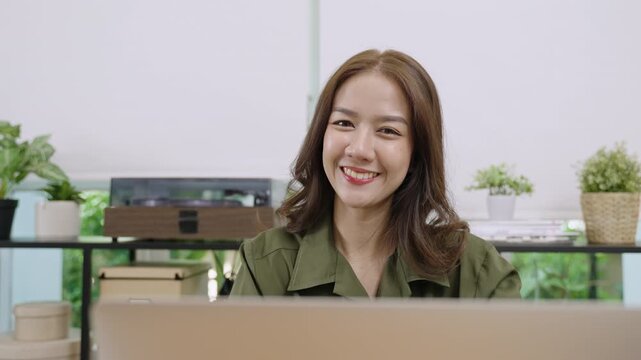Portrait of happy beautiful young Asian business woman sitting at her desk and using a laptop computer for work while looking at the camera and smiling cheerfully at the workplace in the office. 