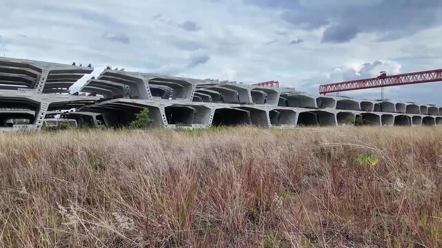 Massive precast concrete bridge segments are neatly stacked in a vast, overgrown grassy field under a cloudy sky.