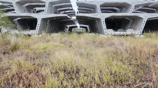 Massive precast concrete bridge segments are neatly stacked in a vast, overgrown grassy field under a cloudy sky.