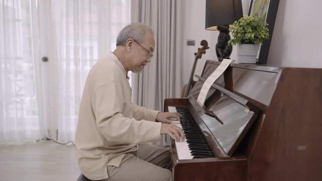 A happy Asian senior man pianist in glasses is sitting, playing classical piano alone after retirement, relaxing in the living room at home. Elderly male playing a keyboard instrument with happiness. 