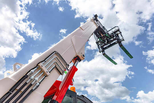 Telescopic boom arm and pallet fork attachment on telehandler machine. Extended hydraulic boom lifter low angle view featuring steel pipes, red cylinder, fork carriage, and cloudy sky.