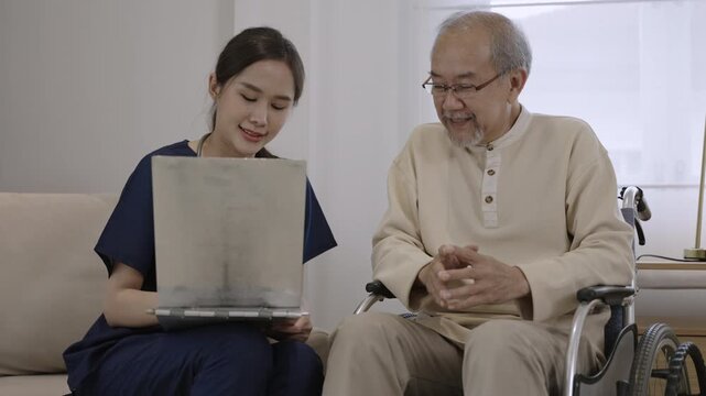 Professional young Asian woman nurse caregiver holding clipboard is talking, explaining, and providing healthcare advice to senior male patient while sitting in wheelchair in special room at hospital.