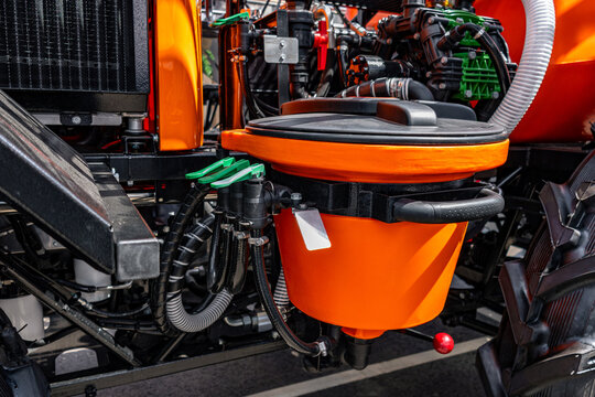 Chemical induction hopper and control valves on field sprayer. Detail of agricultural chemical mixing tank featuring orange bowl, sealed lid, green lever valves, and diaphragm pump behind.