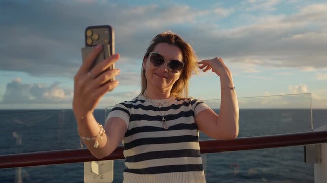 Woman in striped shirt takes selfie photos on cruise liner deck in Caribbean Sea. She poses with smartphone while wind blows her hair against ocean horizon and dramatic clouds during sunset.