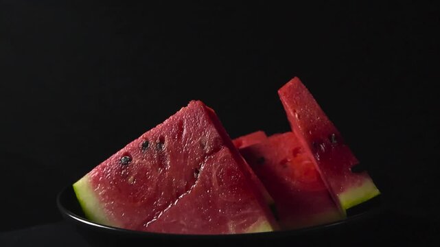 Refreshing triangular slices of ripe, juicy watermelon with seeds slowly rotating on a black plate. The vibrant red pulp and green rind contrast beautifully against the dark, moody background