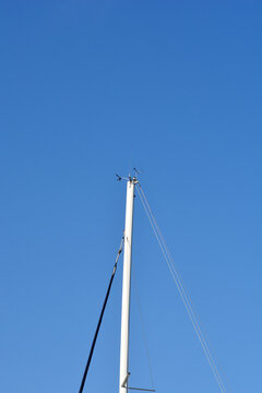 The white masthead of a sailboat features a wind vane anemometer and radio antenna against a solid blue sky background