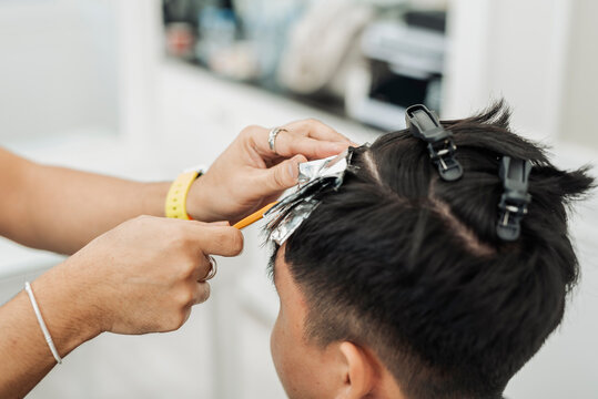 Hair stylist sectioning hair and placing foils for highlights during professional hair highlighting treatment for an Southeast Asian man in his early 20s