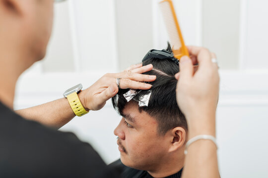 Hair stylist sectioning hair and placing foils for highlights during professional hair highlighting treatment for an Southeast Asian man in his early 20s
