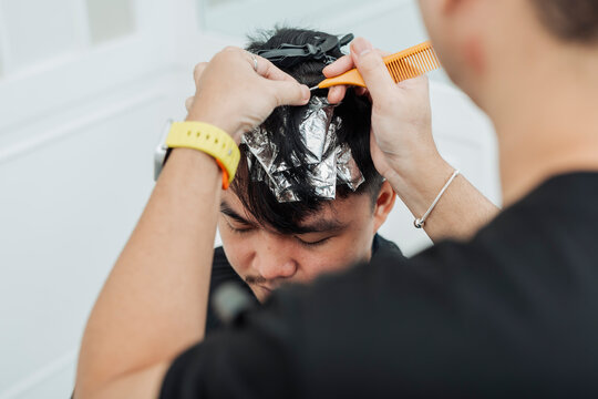 Hair stylist sectioning hair and placing foils for highlights during professional hair highlighting treatment for an Southeast Asian man in his early 20s