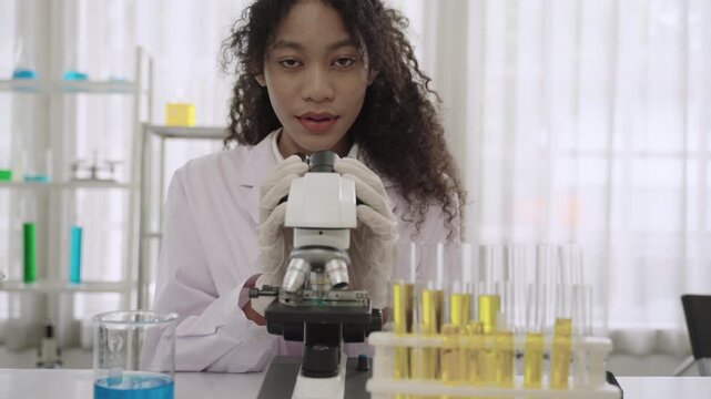 Portrait of a happy professional young woman scientist and researcher in a lab coat wearing safety gloves sitting smiling and using a microscope while looking at the camera in the science laboratory. 