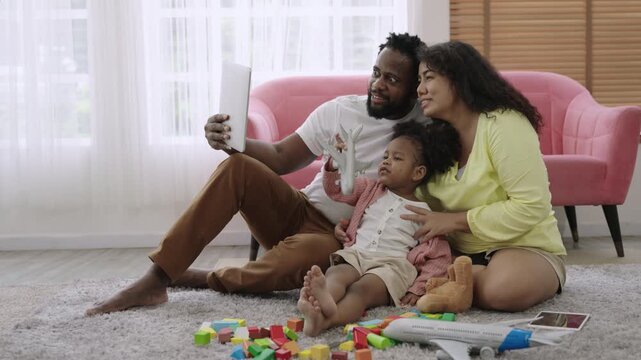 Happy Family Day concept. African American family sitting, smiling, and using a tablet for a video call while their little daughter plays with plane model, with happiness on living room floor at home.