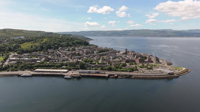 A cinematic aerial orbit showcasing the picturesque coastal town of Gourock, Scotland, featuring the harbor, residential architecture, and the vast Firth of Clyde under a bright blue sky.