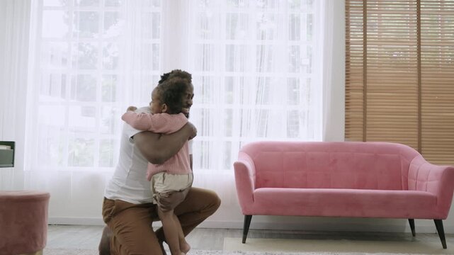 A cute African American child daughter is running and smiling to embrace her father while sitting and waiting on the carpet floor, happy and bonding in the living room at home. Father's Day concept.