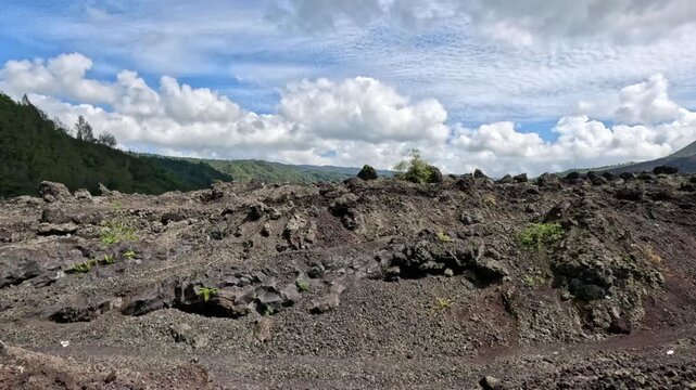 Wide shot of Bali's lava valley showing solidified black lava, green hills, and Mount Batur under white clouds during daytime. Volcanic landscape with tropical vegetation.