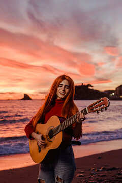 Portrait of redheaded young woman playing guitar on the beach at sunset, Almunecar, Spain