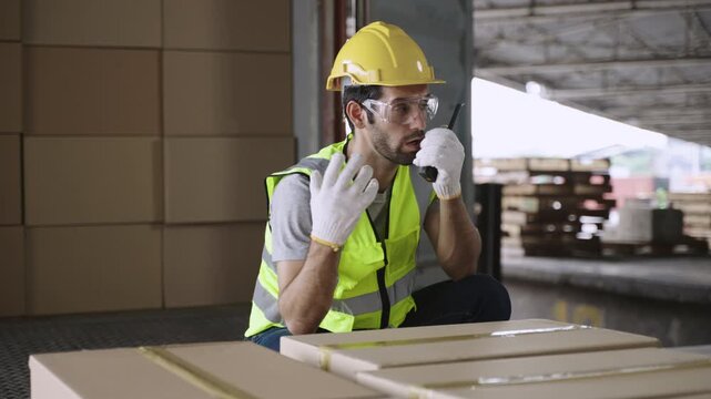 Handsome young man warehouse inspector in a uniform wearing goggles and yellow safety helmet, using a walkie-talkie while talking and discussing while inspecting cargo boxes in a distribution factory.