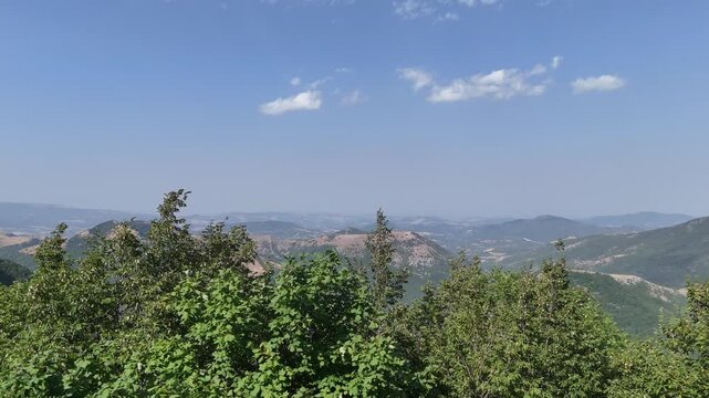 Paesaggio naturale con alberi verdi e vegetazione fitta in primo piano, colline e montagne lontane dai toni azzurri e cielo limpido con leggere nuvole