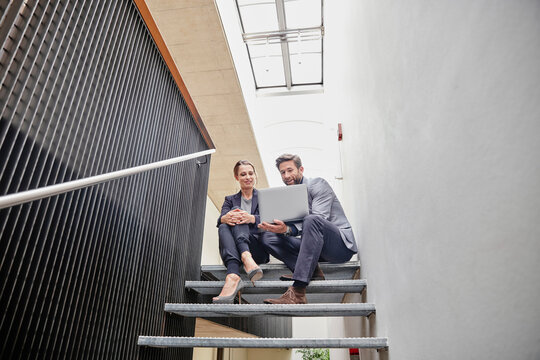 Businessman and businesswoman sitting on stairs in office sharing laptop