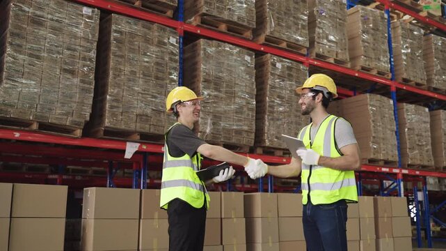 Happy warehouse workers in uniform wearing yellow helmets holding tablet shaking hands, working, talking, discussing and planning together in logistics distribution factory. Inventory stock concept. 