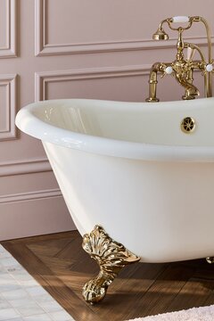 Closeup of clawfoot tub rim in bone white enamel with champagne brass hardware against a matte dusty rose plaster wall in soft morning light