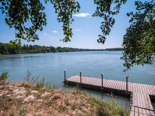 Fototapeta premium Wooden floating dock on the Danube River surrounded by trees, offering peaceful access for boating, kayaking, canoeing and other water activities in a natural riverside landscape.