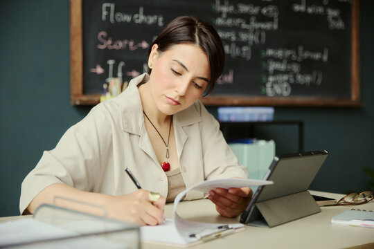 Young adult Caucasian woman reviewing papers and writing notes beside tablet in classroom, studying lesson plan and workflow concepts. Useful for education, training, tutoring, academic content