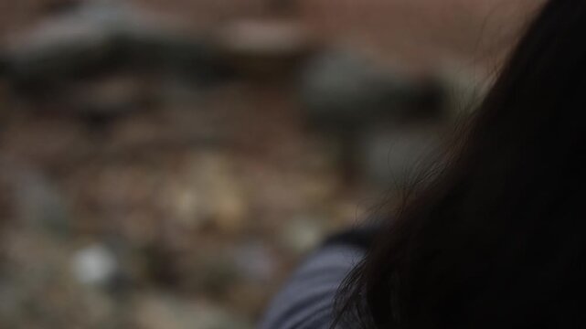 A person in casual clothing is partially visible over the shoulder, positioned next to a rocky outcrop outdoors. Soft, diffused natural lighting with earthy textures sets a contemplative mood.