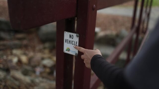 Close-up shot of a person pointing to a metal 'No Vehicle' sign on a rustic wooden bridge, with autumn leaves and a rocky stream in soft, natural daylight. Shallow depth of field.