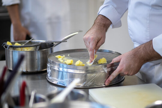 Closeup Professional Chef Hand Pressed Mashing Boiled Potatoes Through Drum Sieve Pot with Plastic Scraper Working in Restaurant Kitchen