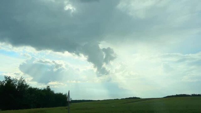Driving past a green field as weather begins to shift. Blue sky with gathering rain clouds and sun rays breaking through. Changeable summer scene captured along a countryside road..