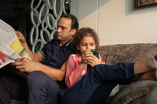 Young girl spending time on the phone while the father reads the newspaper at home. 