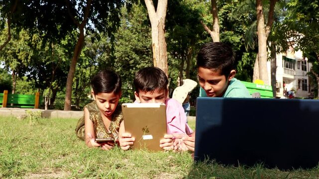 Elementary age cute loving siblings lying down on the grass outdoor in nature during summer season and using Laptop, Digital tablet and mobile phone together video portrait.