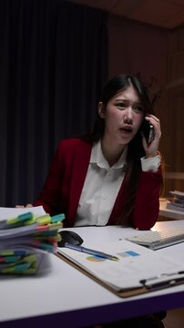 Upset Asian businesswoman talking on her phone getting bad news. A frustrated employee working overtime in a dark office at night.