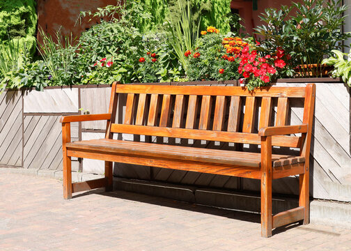 Wooden park bench garden photo. Empty varnished timber seat stands on brick pavement in front of blooming flowerbed with red orange blossoms. Classical outdoor backyard patio furniture design closeup 