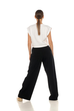 Full length rear view fashion portrait of woman standing with wide stance, white crop top and black trousers, on white background.