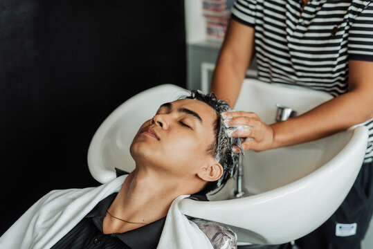 Hair stylist rinsing and washing young man's hair before professional haircut in salon for Southeast Asian man in his early 20s
