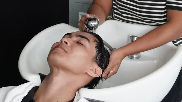 Hair stylist rinsing and washing young man's hair before professional haircut in salon for Southeast Asian man in his early 20s