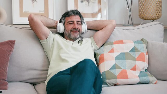 Video of mature man listening music with headphones while relaxing on couch at home