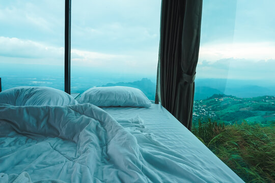 A cozy, unmade bed showcasing a breathtaking panoramic mountain view through a large floor-to-ceiling glass window at a luxury highland resort. 