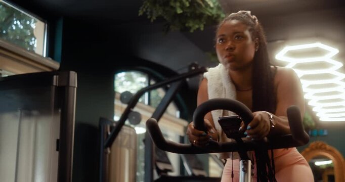 Focused Black woman rides a stationary bike during an indoor cardio workout in a modern gym.