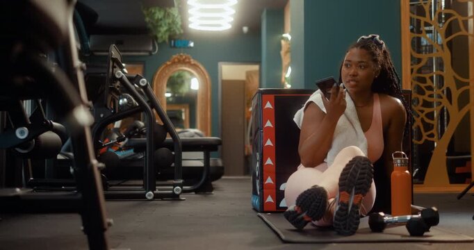 Black woman rests on the gym floor during a workout break while recording a voice message on her smartphone.