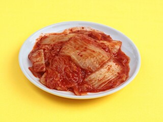Fototapeta premium High angle view Korean kimchi cabbage served in white plate isolated on yellow background. Ideal for Asian food concepts, healthy eating, probiotics, and traditional Korean cuisine