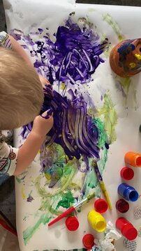 Young Child Making Messy Purple Finger Painting, Overhead View - Vertical overhead footage of a young child pouring and spreading thick purple paint across paper with paint-covered hands during a colo