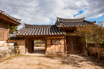Fototapeta premium Exterior View of Traditional Korean Hanok Entrance Gate Featuring Courtyard, Stone Wall, and Tiled Roofs Under Cloudy Sky