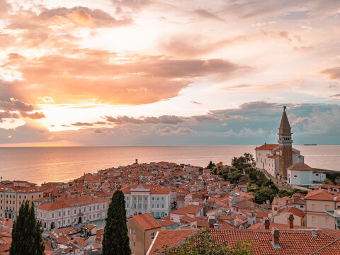 A picturesque view of Piran, a coastal town with red-tiled roofs and a prominent church tower is bathed in the warm glow of a sunset over the calm sea
