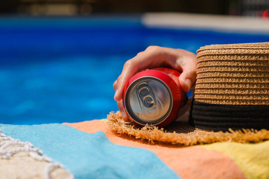A can of cold drink by the pool. A hand reaches for the drink. Summer relaxation.