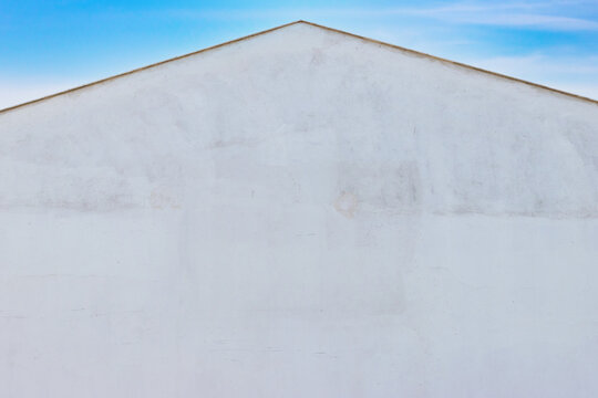 Minimalist off-white textured stucco wall with a pitched gable roofline and subtle stains, set against a clear bright blue sky for architectural background use.